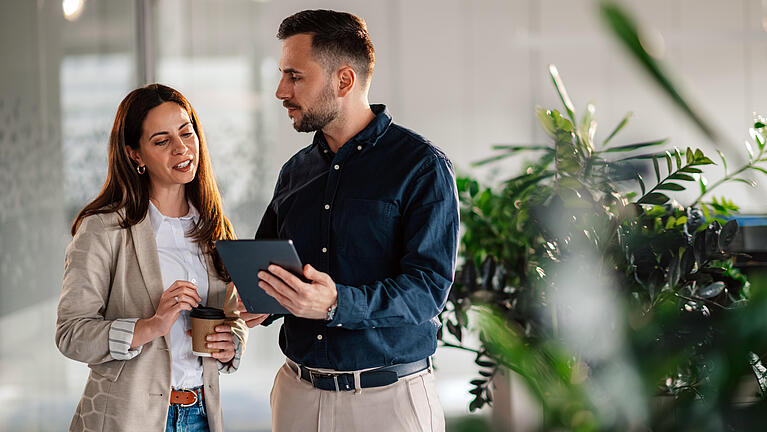 Frau in hellem Blazer spricht mit Mann im Büro über Angebote auf iPad.