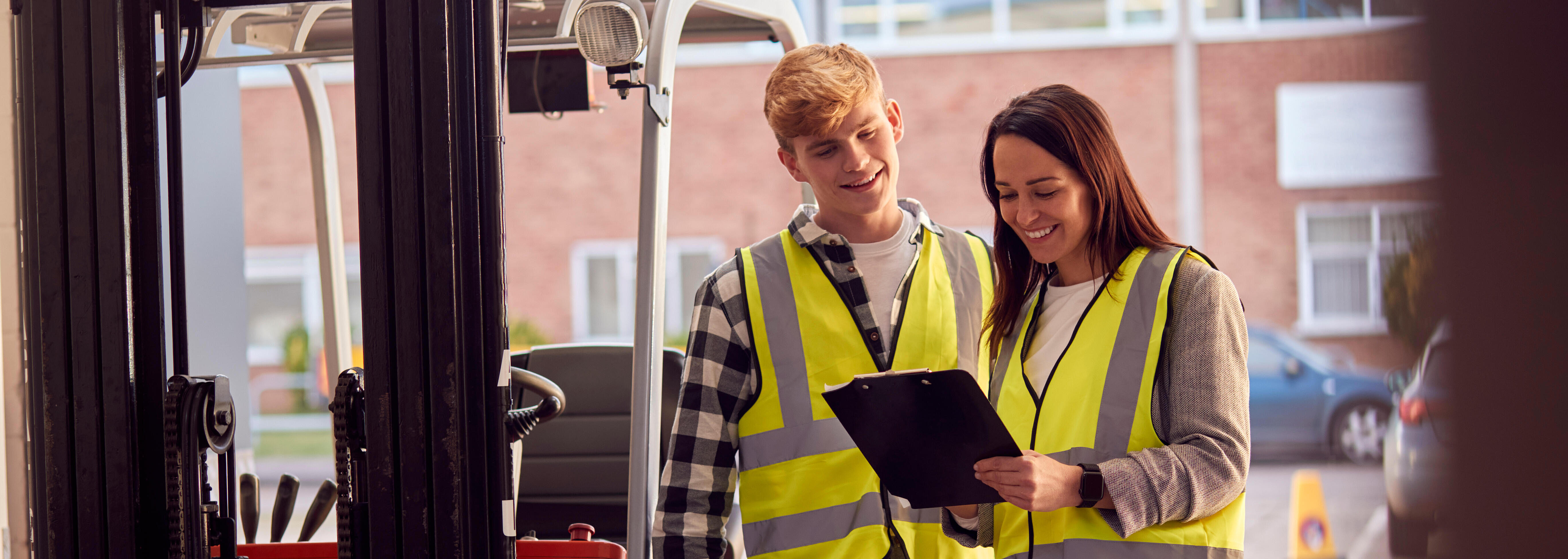 Auszubildender und Ausbilderin in Warnwesten auf einem Betriebsgelände vor einem Gabelstapler